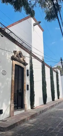 White stucco building on a cobblestone street, ornate arched doorway with a carved crest above and tall slender trees along the wall under a blue sky, San Miguel de Allende