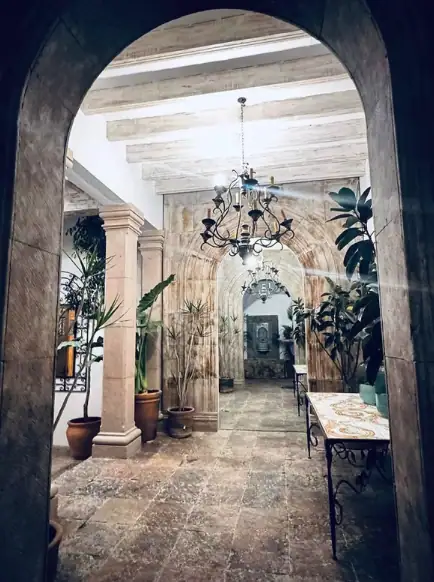 Stone arched doorway into a plant-filled hallway with a chandelier and a table on the right, San Miguel de Allende