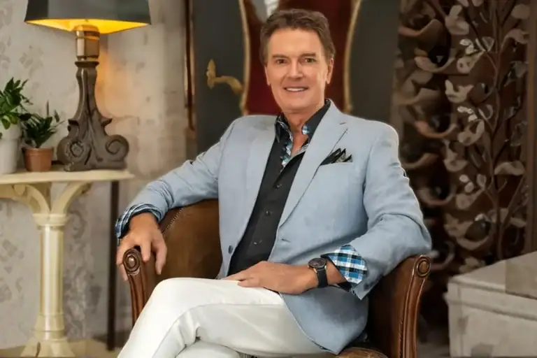 Greg Gunter seated in a leather armchair in a light blue blazer and white trousers beside a lamp and side table