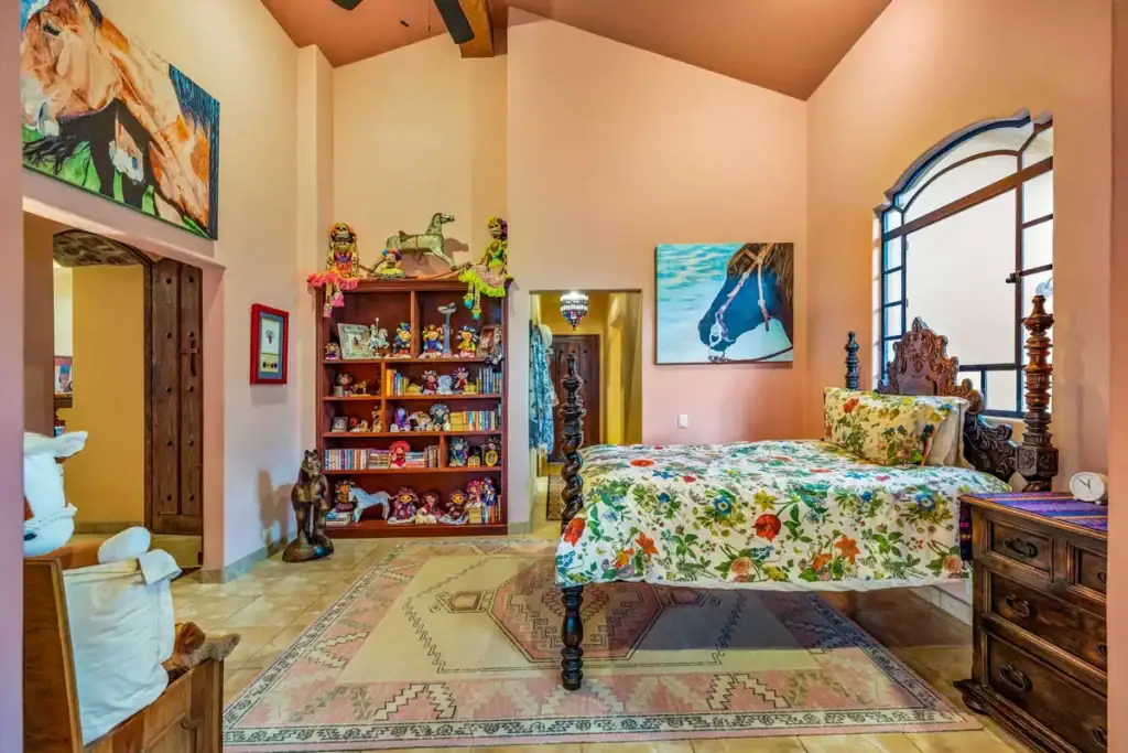 Bedroom with a carved wooden bed, bookshelf of folk-art figures and a large arched window, San Miguel de Allende