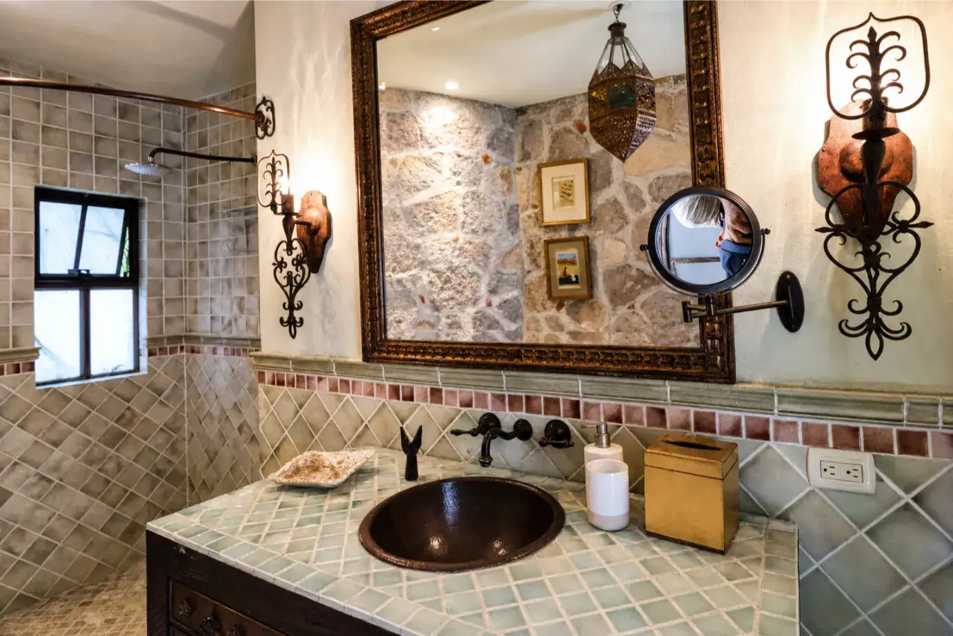 Rustic bathroom with a tiled vanity, dark copper sink, and an ornate framed mirror with vintage lighting fixtures on either side, San Miguel de Allende