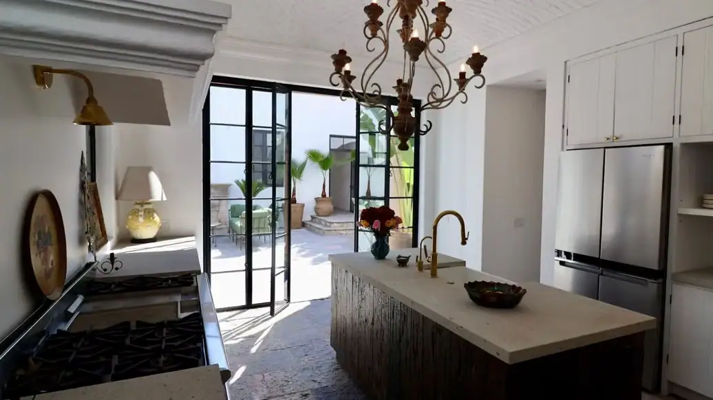 Bright modern kitchen with a large island, brass faucet, and a chandelier, opening to a sunlit outdoor patio through black-framed glass doors, San Miguel de Allende
