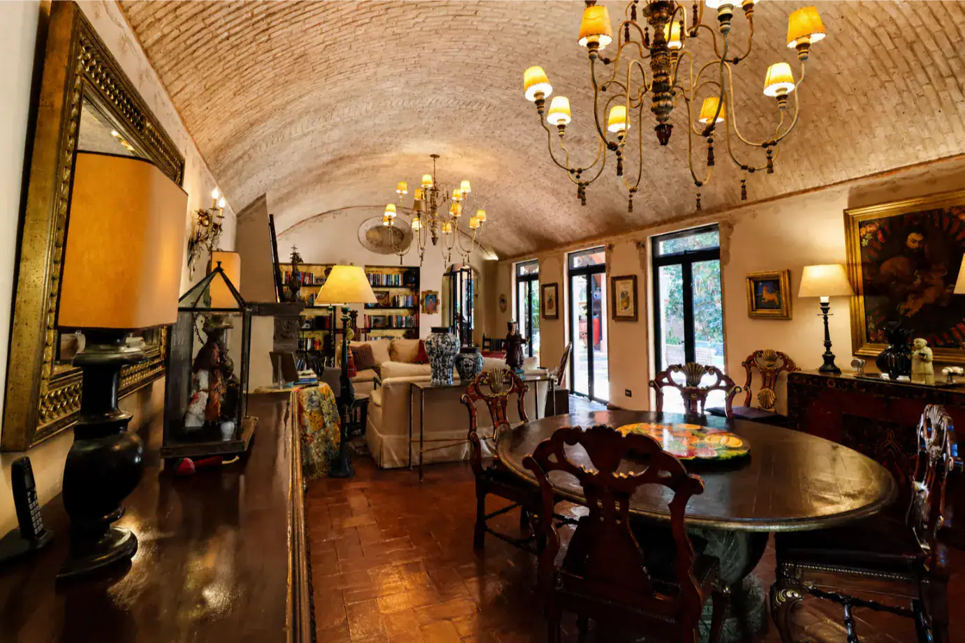 Cozy living and dining room with a brick vaulted ceiling, ornate chandeliers, a round wooden table, and bookshelves lining the back wall, San Miguel de Allende