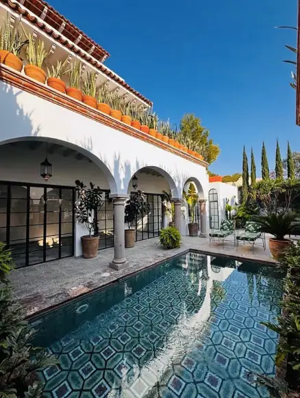 A white building with arched colonnade and a tiled pool along the courtyard, lined with potted plants and a blue sky above, San Miguel de Allende