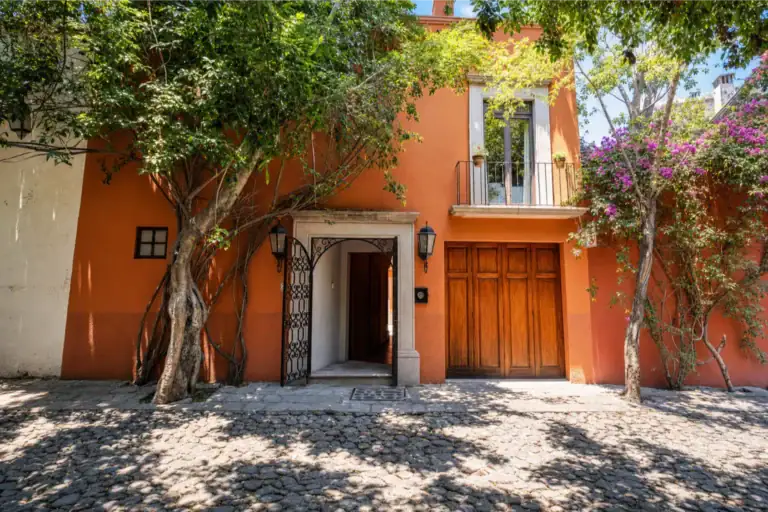 Two-story orange building with a wooden double door and iron gate, framed by lush vines on a cobblestone street. The upper balcony has potted plants and yellow railing accents, San Miguel de Allende
