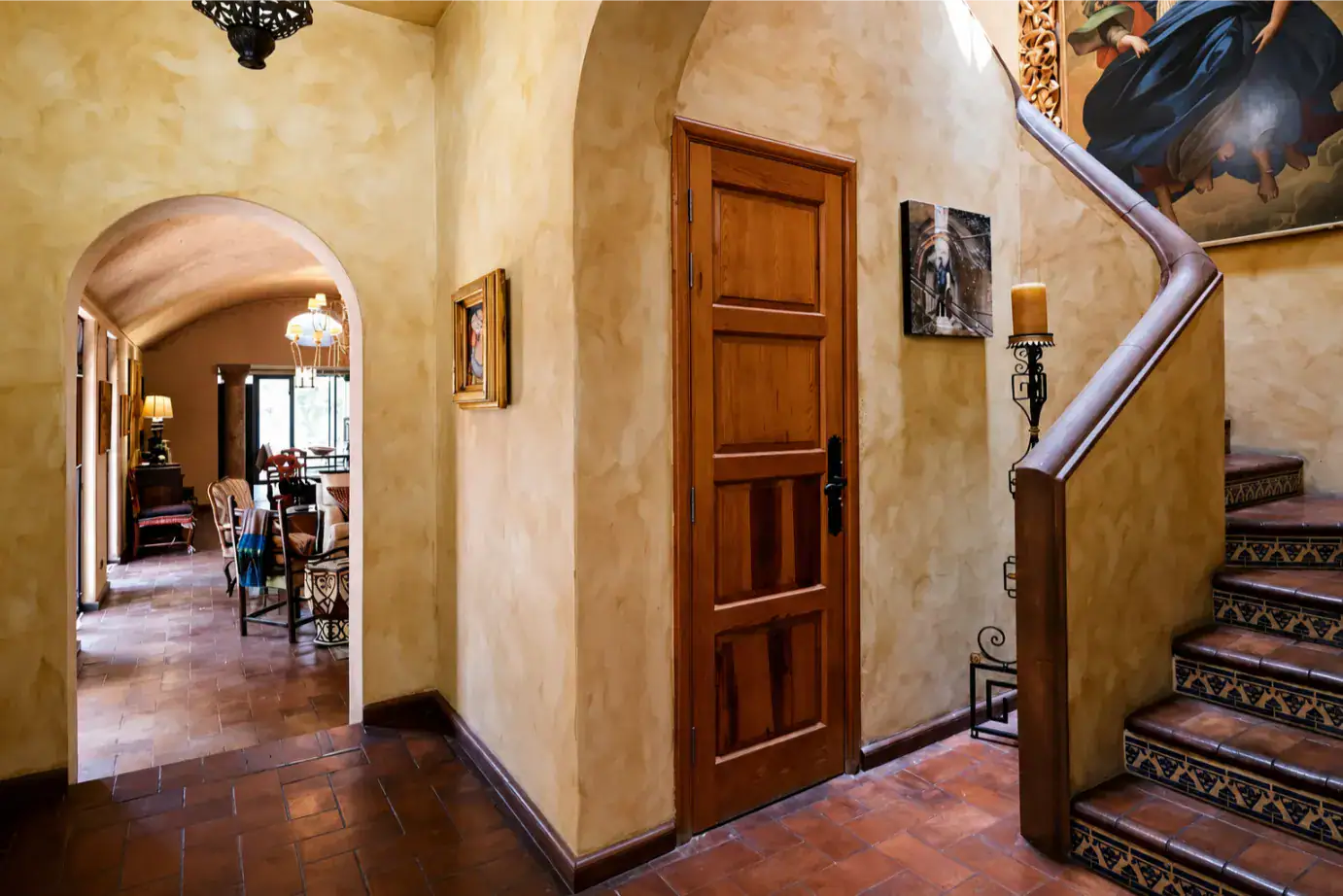 Warm Mediterranean-style entry hall with a wooden door, arched doorway to a living room, and a tiled staircase railing, San Miguel de Allende