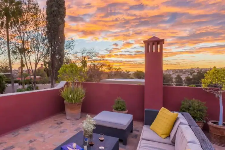 Rooftop terrace with a red-walled chimney, outdoor sofa and potted plants under a sunset sky in San Miguel de Allende