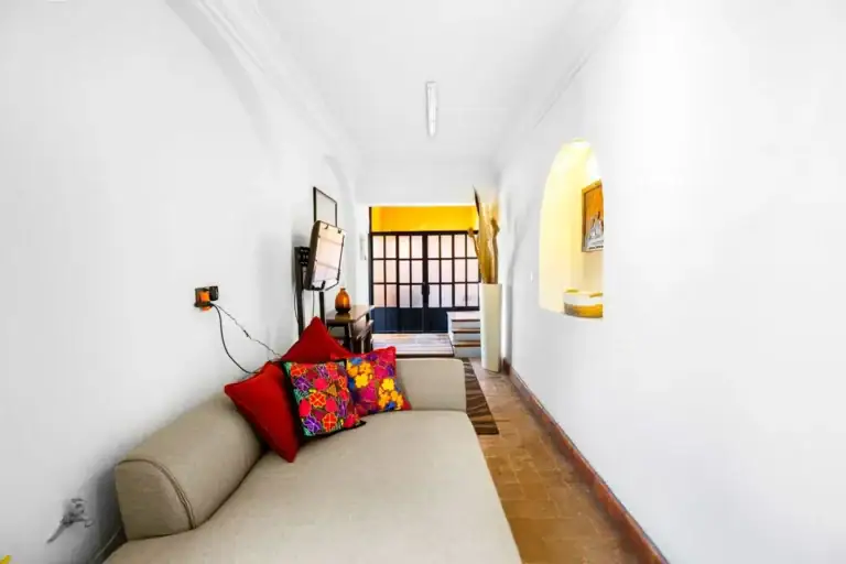 Santo Domingo House, narrow sitting room with a beige sofa, a red embroidered cushion and a view to the next room, San Miguel de Allende