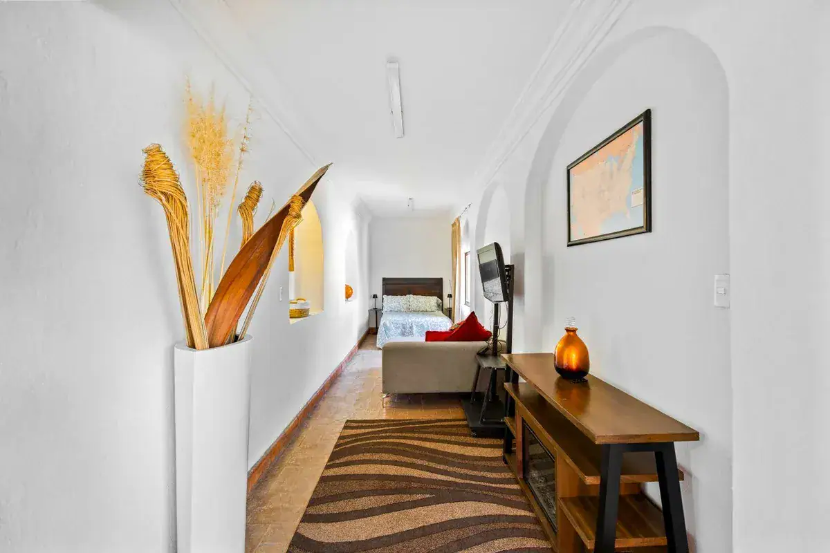 Santo Domingo House, hallway with a tall white vase of dried reeds, a bedroom beyond and a patterned rug, San Miguel de Allende