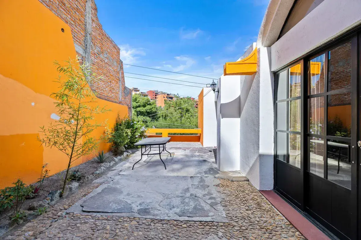 Santo Domingo House, stone patio with a bistro table, an orange wall and a tree beside black-framed glass doors, San Miguel de Allende