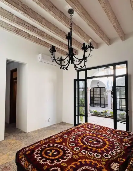 Bedroom with a black ornate chandelier, exposed wooden ceiling beams, and a vibrant patterned bedspread; double glass doors open to an outdoor courtyard, San Miguel de Allende