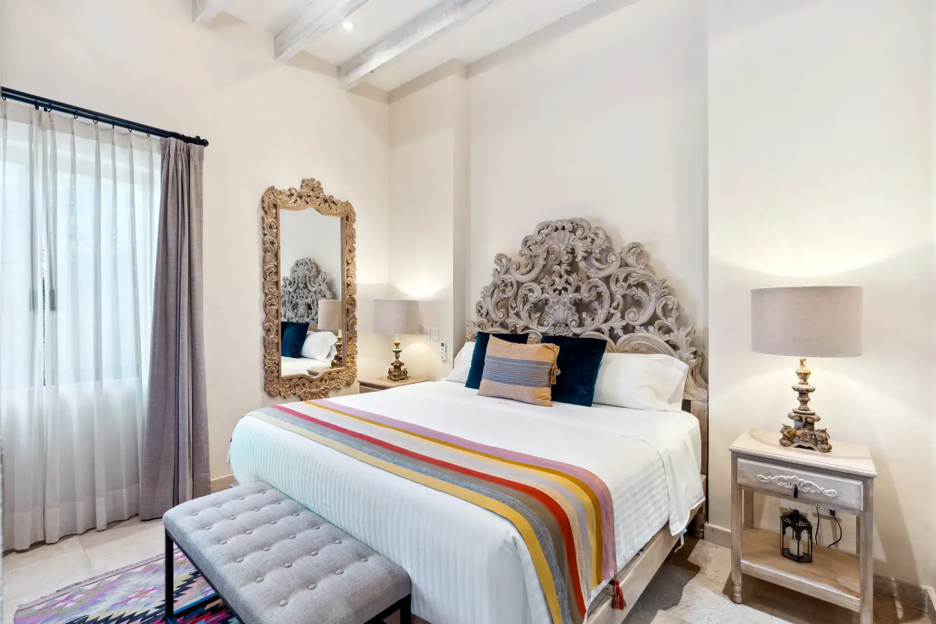Suites Casa Vistas, bedroom with a carved gray headboard, striped blanket and a tufted bench, San Miguel de Allende