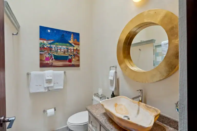 Suites Casa Vistas, bathroom with a stone vessel sink, round gold mirror and a colorful painting of a town square, San Miguel de Allende