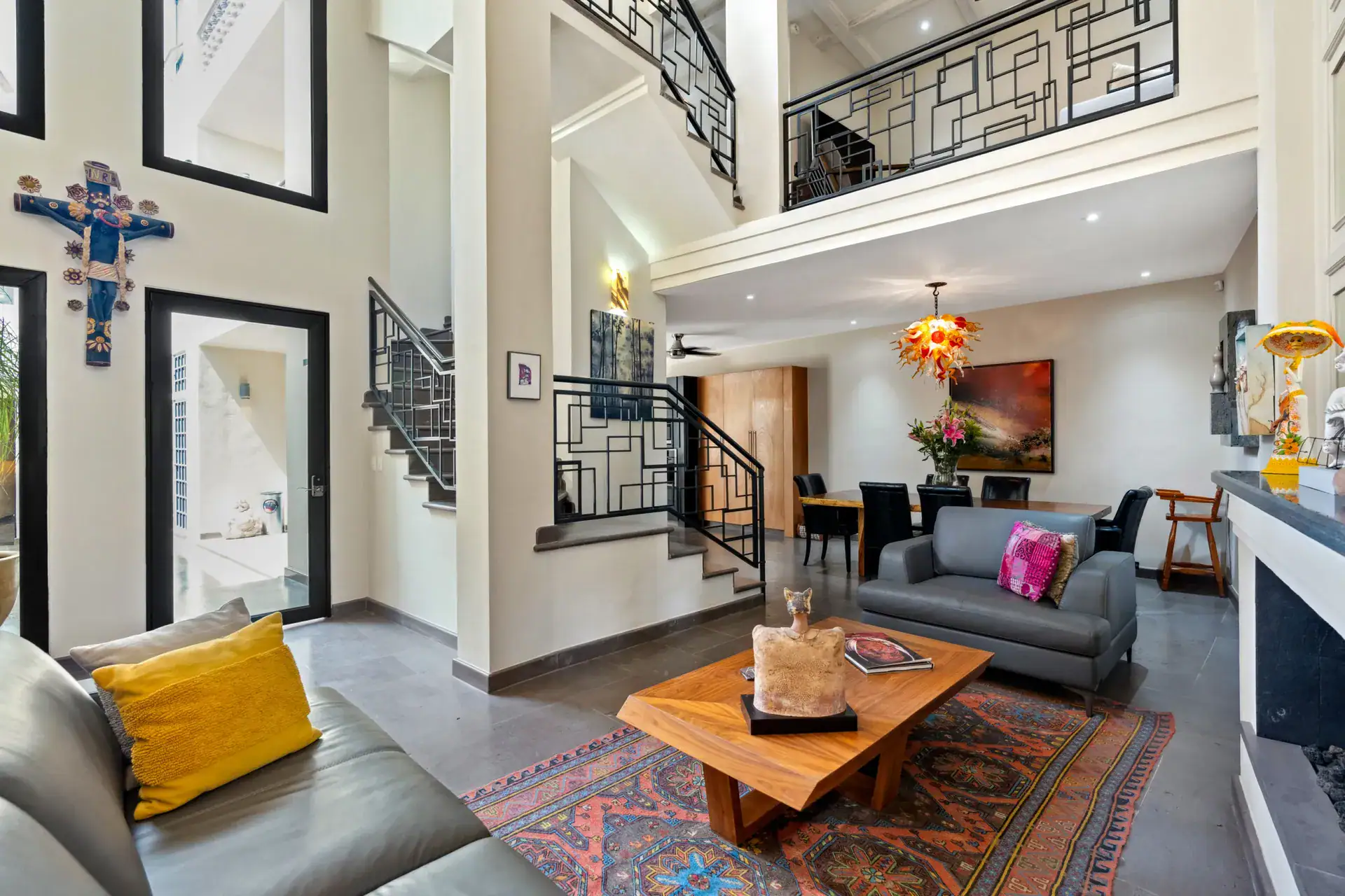 Suites Casa Vistas, double-height living room with a gray sofa, wood coffee table, wrought-iron stair rail and a fireplace, San Miguel de Allende