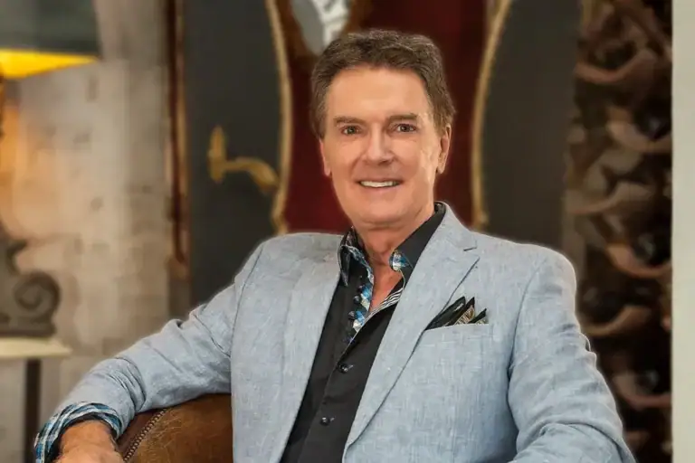 Smiling man in a light blue blazer seated with a guitar in a stylish indoor setting.