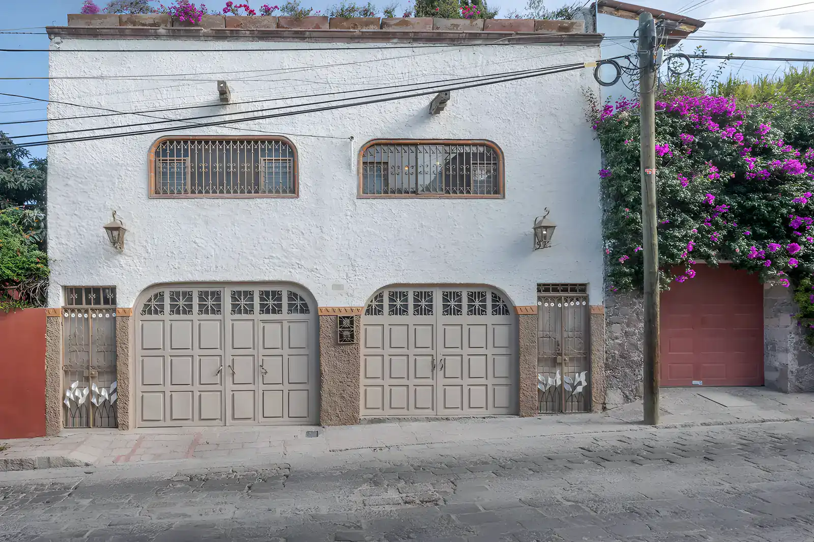 Two Homes, white stucco street facade with two paneled garage doors, barred arched windows and a pink bougainvillea, San Miguel de Allende