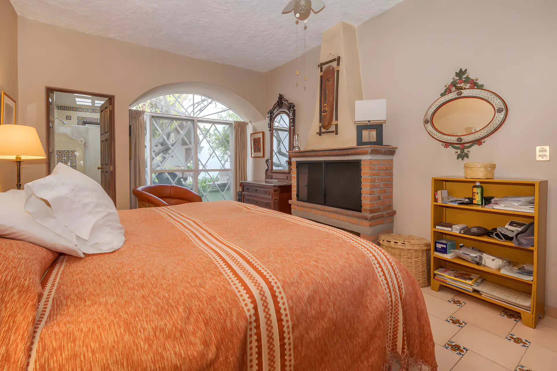 Two Homes, bedroom with an orange bedspread, a brick fireplace, an arched window and a bookshelf, San Miguel de Allende