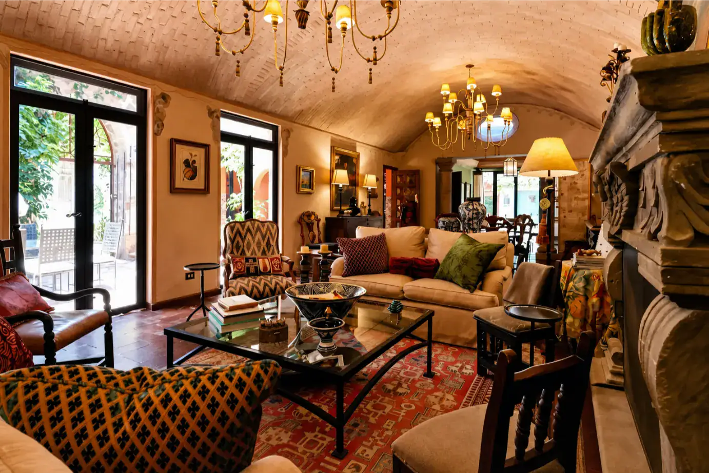 Cozy living room with warm lighting, a cream sofa, patterned chairs, and a glass coffee table beside a stone fireplace under vaulted ceilings, San Miguel de Allende