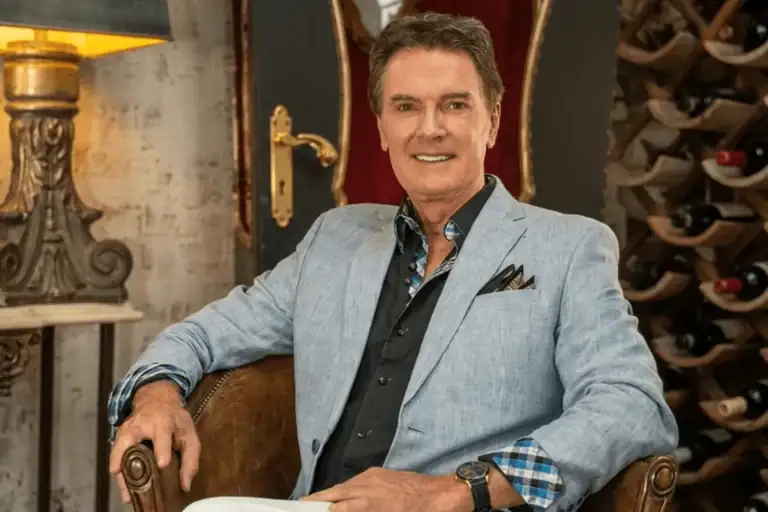 Greg Gunter seated in a leather armchair in a light blue blazer beside an ornate lamp and a wine rack
