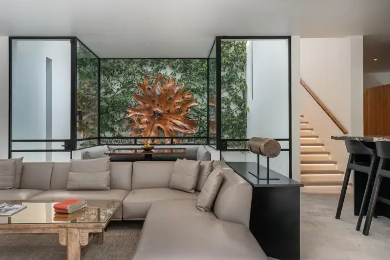 Villa del Parque Central, living room with a large beige sectional, carved wood sculpture, floating staircase and garden glass wall, San Miguel de Allende