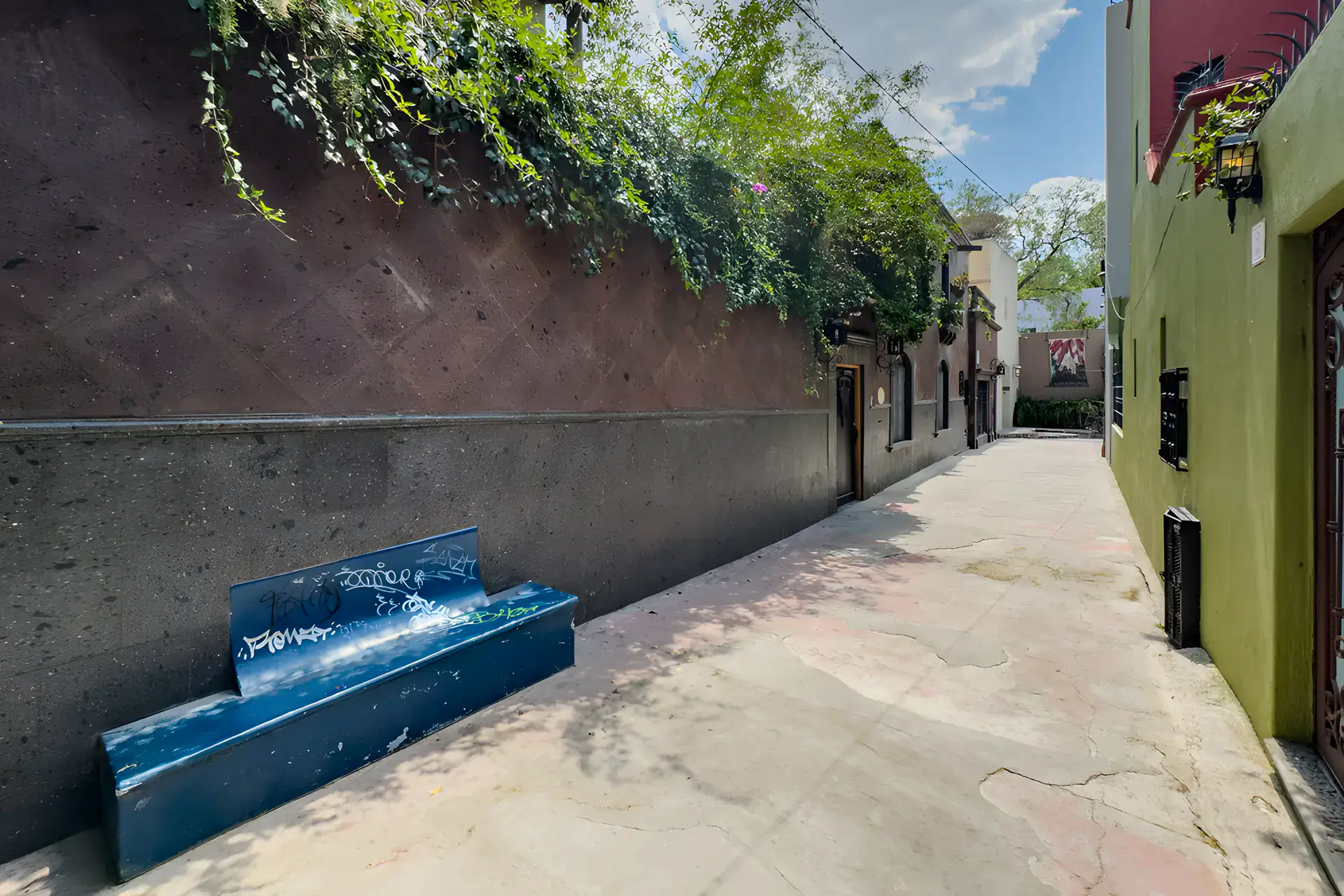 Villa Escondida, narrow street beside a dark stone wall with climbing plants and a blue bench, San Miguel de Allende