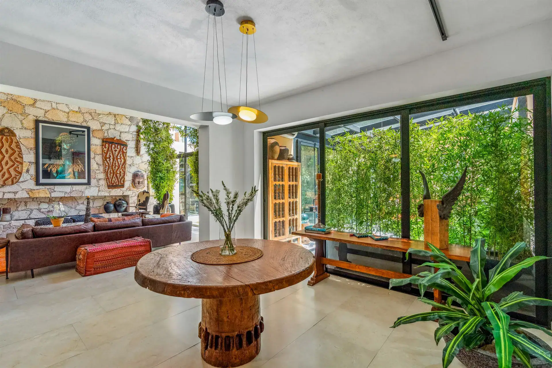 Villa Pacifica, round wood dining table in front of a stone wall, a leather sofa and a glass wall to the garden, San Miguel de Allende
