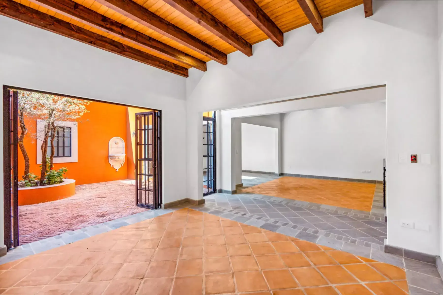 Villa Picasso, empty open room with terracotta and slate floors, a wood beam ceiling and a folding glass wall to an orange courtyard, San Miguel de Allende