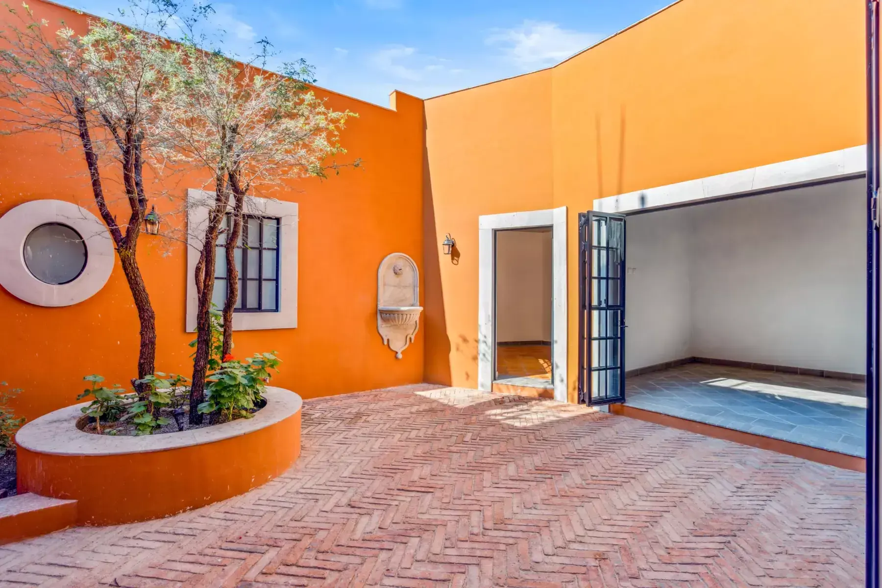 Villa Picasso, orange courtyard with a round planter with trees, a herringbone brick floor and a folding glass wall, San Miguel de Allende