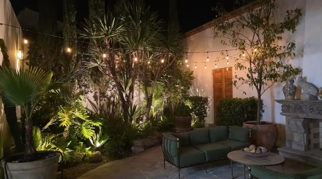 Nighttime courtyard with string lights, tropical plants, and a green outdoor sofa around a circular stone table, San Miguel de Allende