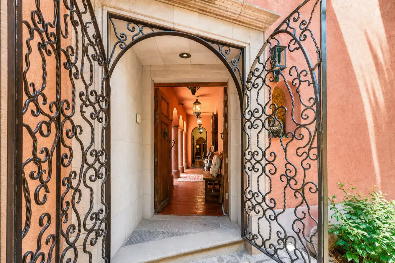 Wrought-iron gates open to a sunlit hallway with arched doorways, warm terracotta walls, and hanging lanterns along a tiled floor, San Miguel de Allende